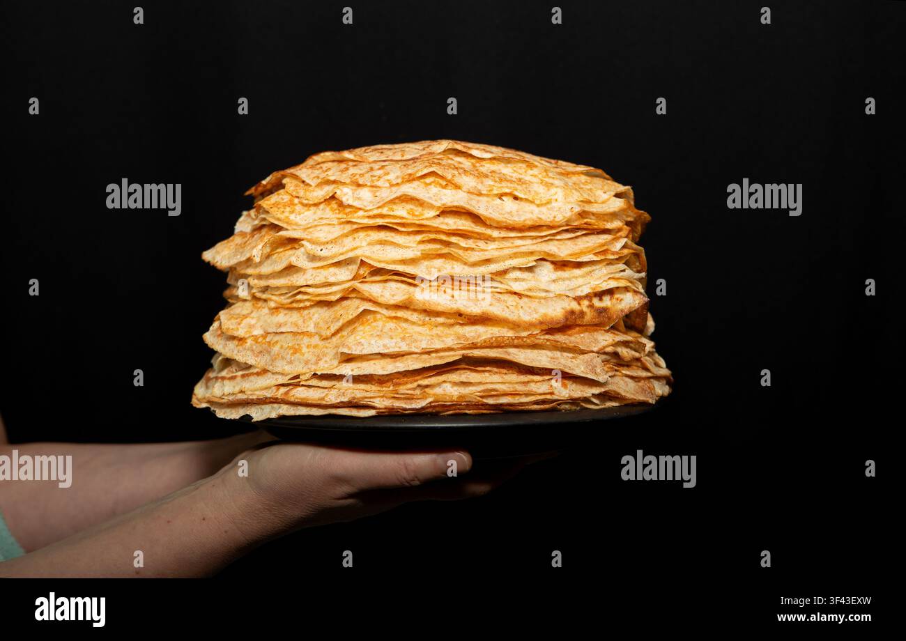 Primo piano di mani che tengono un enorme mucchio di crepe tradizionali fatte in casa su un piatto contro uno sfondo nero scuro. Foto Stock