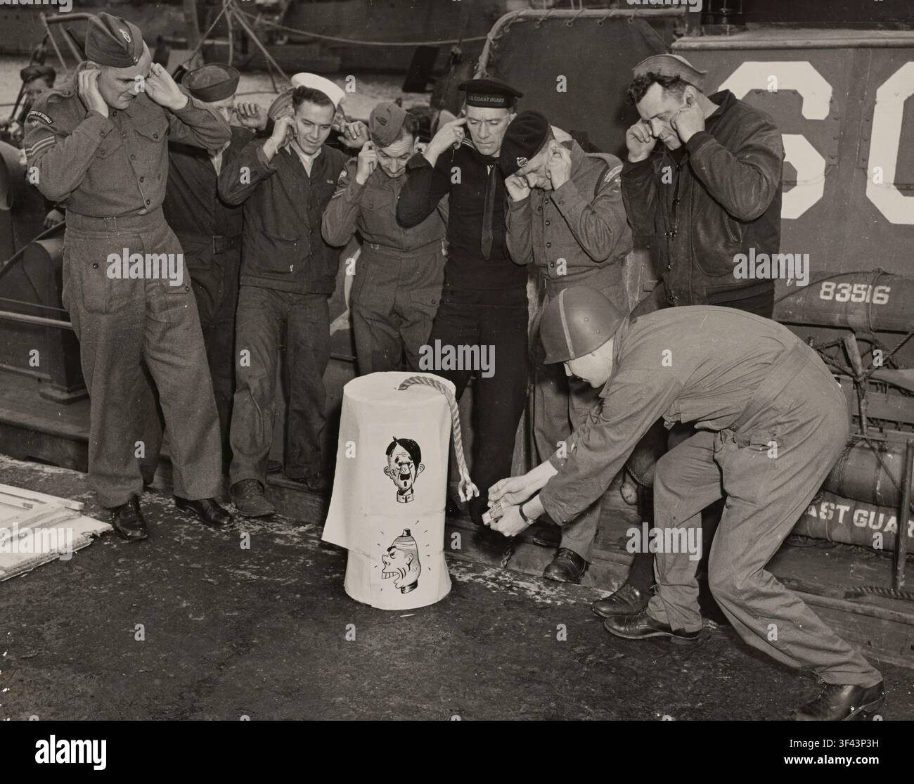 Da qualche parte in Inghilterra, 1944. Membri della Guardia Costiera e soldati dell'esercito britannico si tappano le orecchie mentre un membro della Guardia Costiera accende con un fiammifero la miccia di un petardo sovradimensionato, segnando il Quattro Luglio lontano da casa. Caricature di Hitler e di un leader giapponese in tempo di guerra sono state incise nell'esplosivo gigante per dare all'esplosione festiva una soddisfazione simbolica aggiuntiva e un umorismo benvenuto. Il momento spensierato, inscenato sul ponte di una nave della Guardia Costiera, riflette la facile cameratismo tra le forze americane e britanniche stazionate insieme in vista dell'invasione della Normandia. Foto Stock