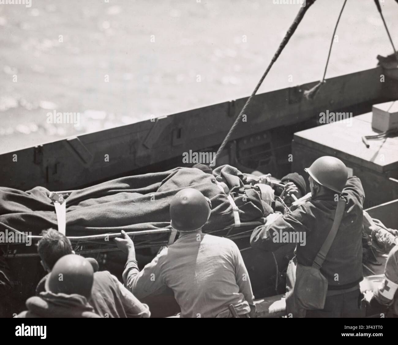 I membri dell'equipaggio di un trasporto d'assalto della Guardia Costiera statunitense trasferiscono con cura i feriti alleati da una nave da sbarco sulla loro imbarcazione al largo della costa francese durante l'invasione della Normandia. Un ferito su una barella avvolto in una coperta, fissato saldamente per il sollevamento, viene guidato a bordo da membri dell'equipaggio caschiati che lavorano sotto una gru e delle sartie, mentre un bracciale della Croce Rossa segna la presenza di personale medico nelle vicinanze. Scene come questa si sono svolte ripetutamente nelle ore successive agli sbarchi del D-Day, mentre le navi al largo lavoravano per evacuare le prime vittime dell'invasione, giugno 1944. Foto Stock
