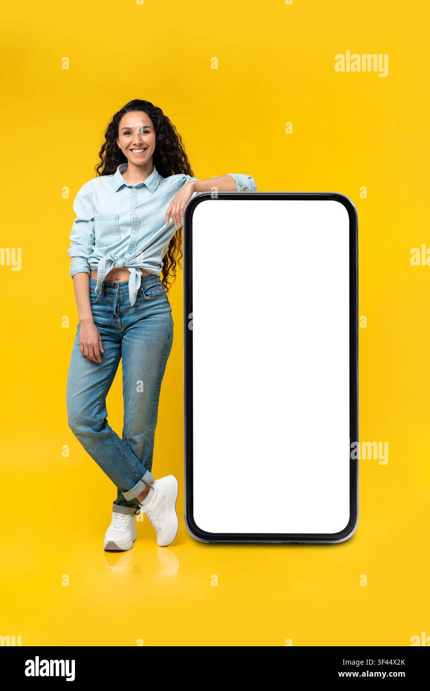 Giovane donna ispanica sorridente appoggiata a uno smartphone con schermo vuoto, sfondo giallo Foto Stock