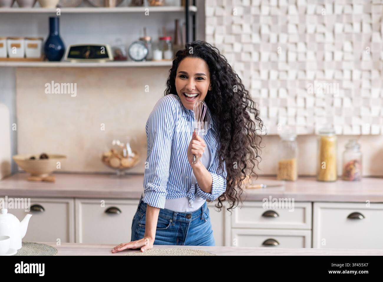 Giovane donna sorridente con capelli ricci che tiene una frusta in una cucina moderna, concetto di cucina Foto Stock