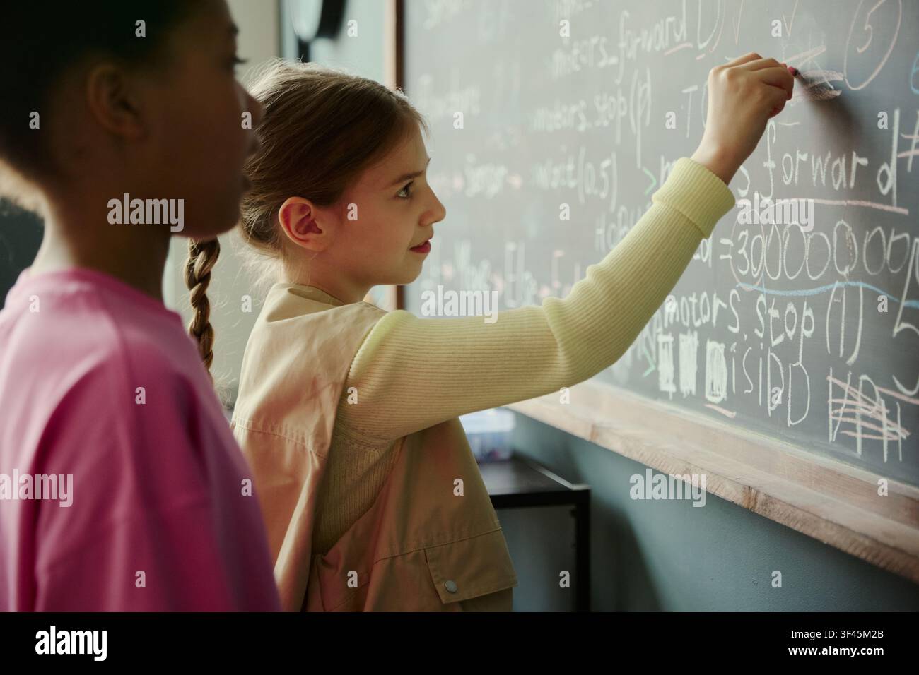 Studentessa caucasica che scrive codice sulla lavagna in classe mentre un compagno di classe nero osserva, mostrando l'apprendimento precoce delle STEM e il lavoro di squadra. Ideale per campagne di educazione, programmazione e tecnologia Foto Stock