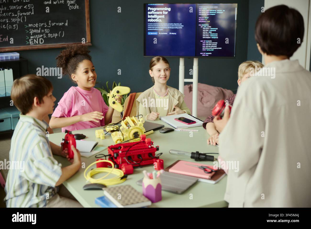Bambini scolastici che imparano robotica e programmazione con l'insegnante, tengono i controller e discutono del progetto al tavolo dell'aula. Ideale per l'educazione STEM, EdTech, promozione del lavoro di squadra Foto Stock