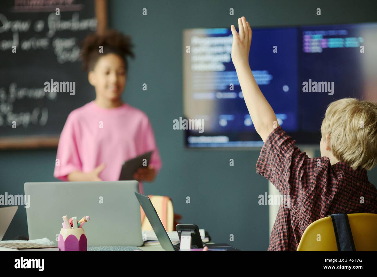 Ragazzino che alza la mano durante la lezione di programmazione mentre una compagna di classe presenta vicino a uno schermo digitale, mostrando il coinvolgimento in classe. Ideale per l'educazione STEM, edtech, corsi di programmazione Foto Stock