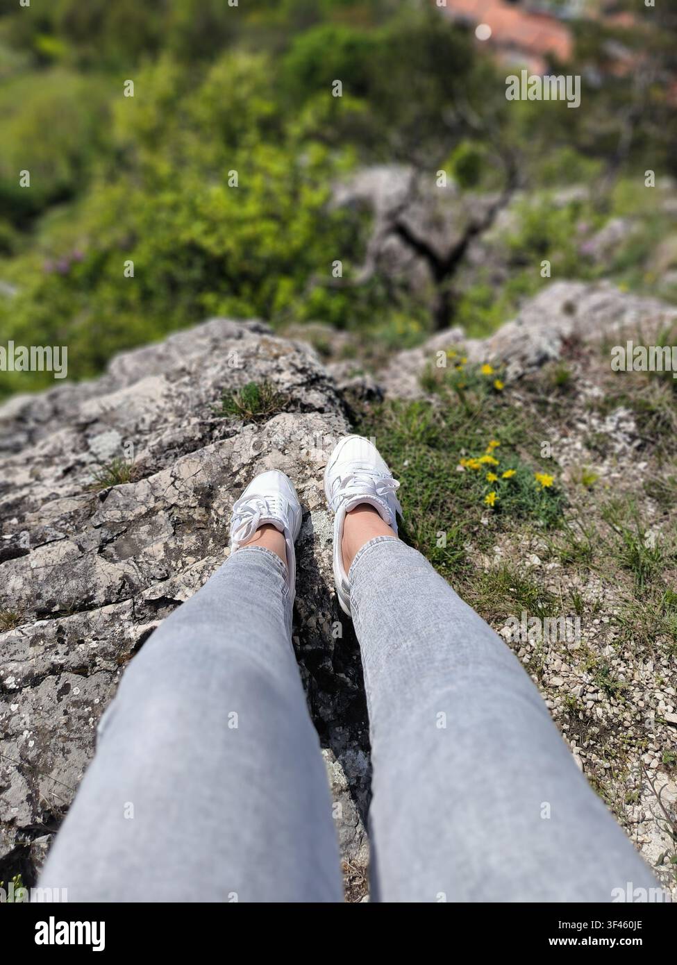 Vista in prima persona delle gambe di una donna in jeans e scarpe da ginnastica seduta sul bordo di una scogliera rocciosa - Immagine stock catturata con smartphone