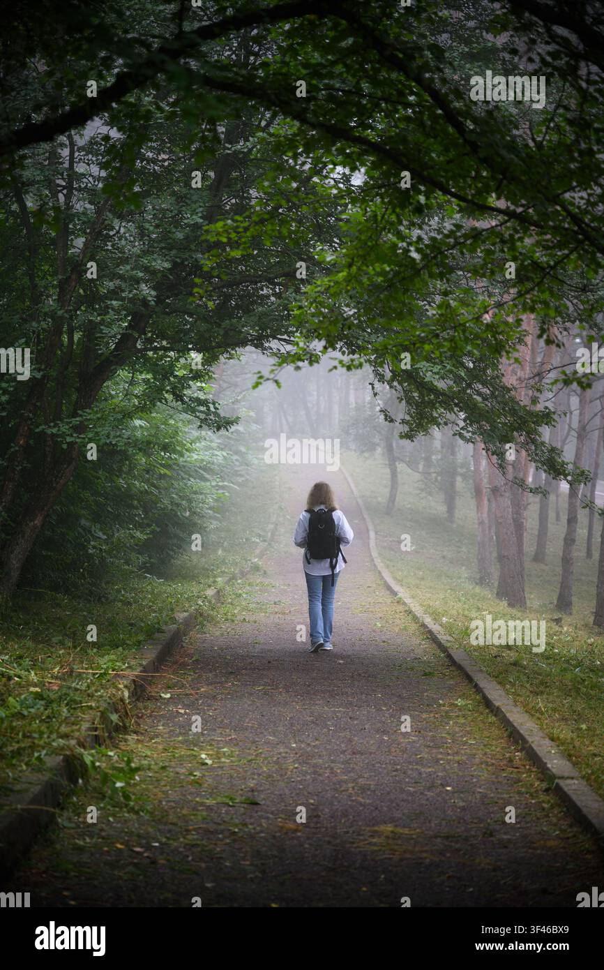 Una foto verticale che mostra una donna che si allontana dalla fotocamera lungo un sentiero lastricato attraverso una fitta foresta. La donna indossa un abito di colore chiaro Foto Stock