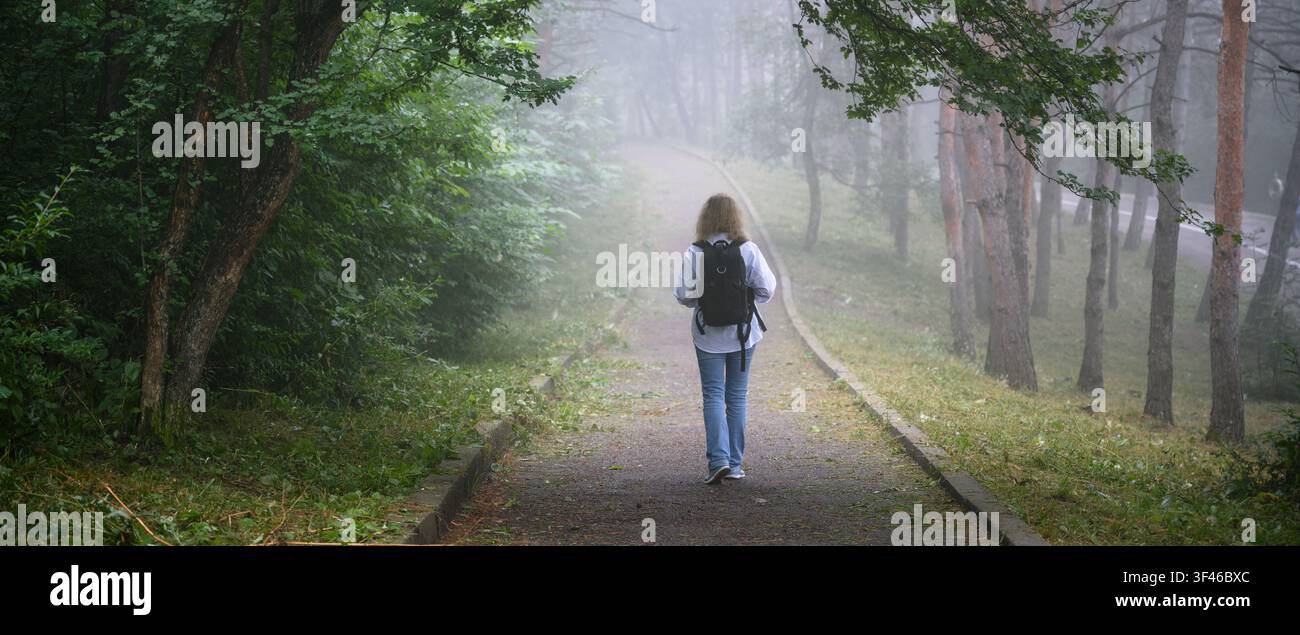 Una fotografia orizzontale di una donna che si allontana dall'osservatore su un sentiero lastricato attraverso una foresta nebbiosa. È vestita in modo casual con blue jeans e un l Foto Stock