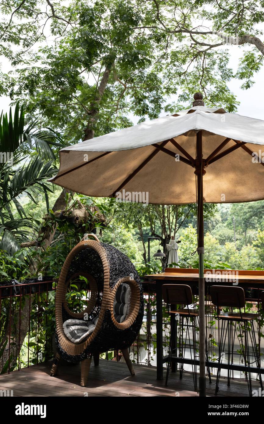 Accogliente sedia a uovo in vimini con ombrellone su terrazza in legno del caffè con vista su una rigogliosa foresta di montagna verde, area seduta all'aperto Foto Stock