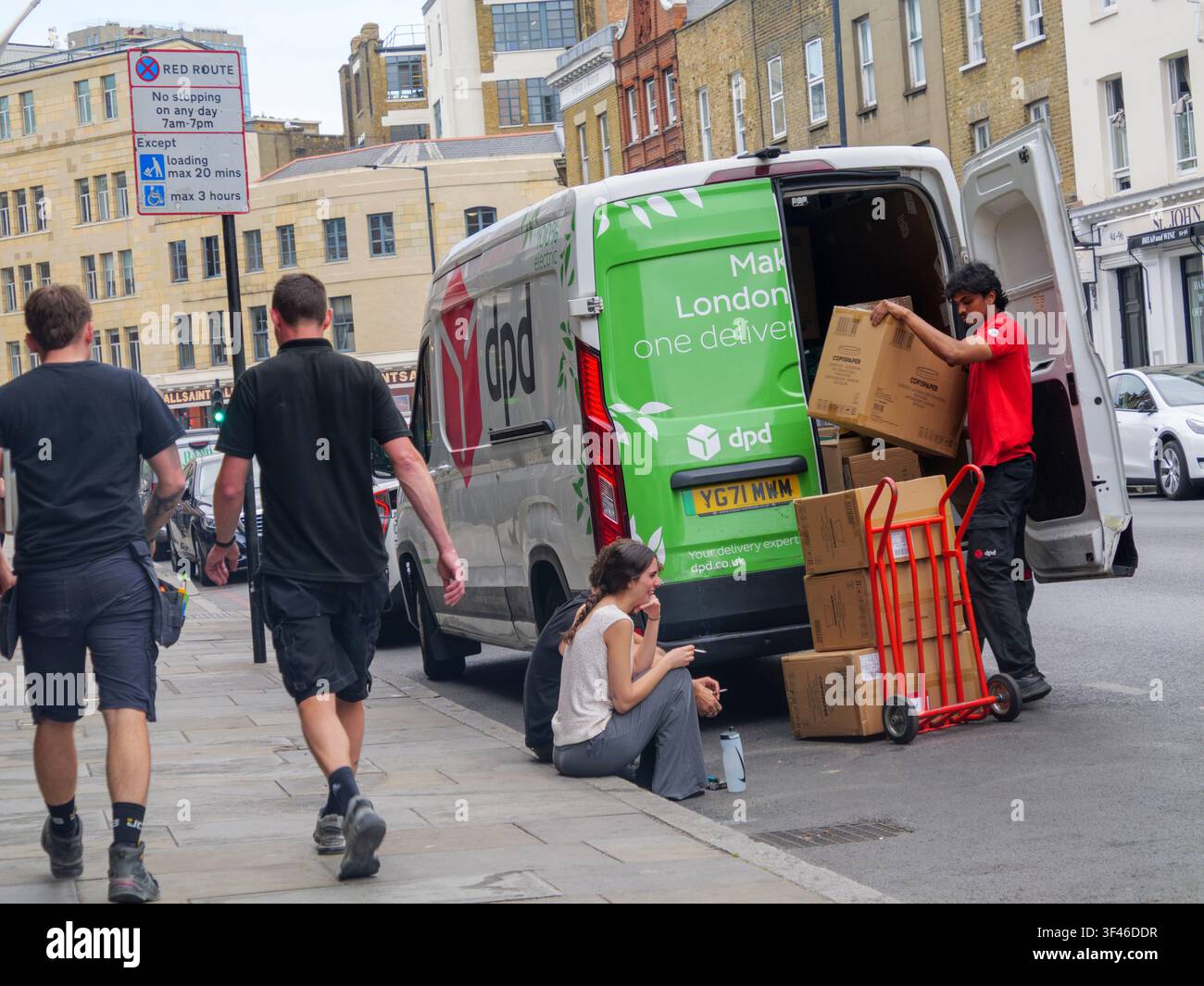 Un autista di consegna DPD scarica pacchi da un furgone di corriere in una strada della City of London, UK, mentre due persone sono sedute sul marciapiede in primo piano prendendo una pausa per fumare una sigaretta Foto Stock