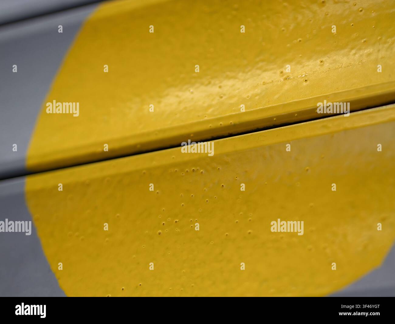Primo piano di una superficie metallica dipinta di giallo con texture, piccole bolle superficiali e uno spazio evidente tra i pannelli su sfondo grigio. Foto Stock
