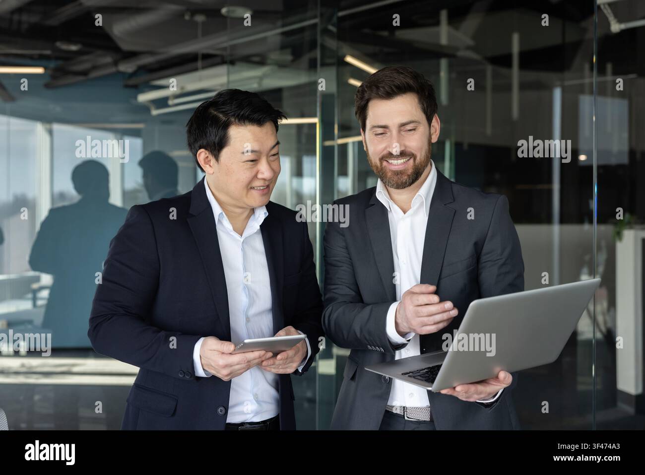 Due uomini d'affari diversificati in piedi in un ufficio moderno, sorridenti e discutendo attivamente del lavoro, interagendo con un laptop e un tablet digitale, dimostrando lavoro di squadra e collaborazione professionale Foto Stock