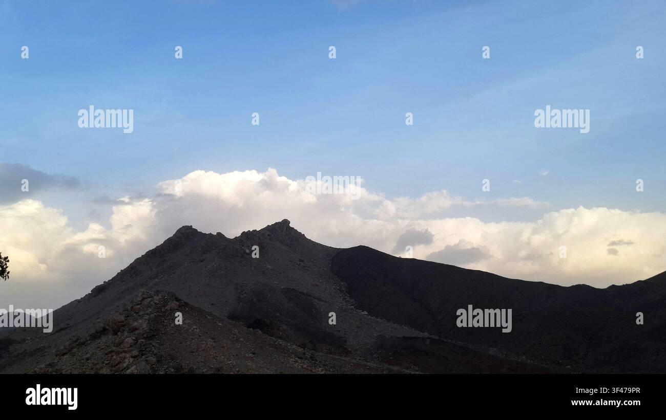 Vista panoramica di picchi montuosi rocciosi e frastagliati sotto un cielo blu chiaro con nuvole bianche in Oman - Immagine stock catturata con smartphone