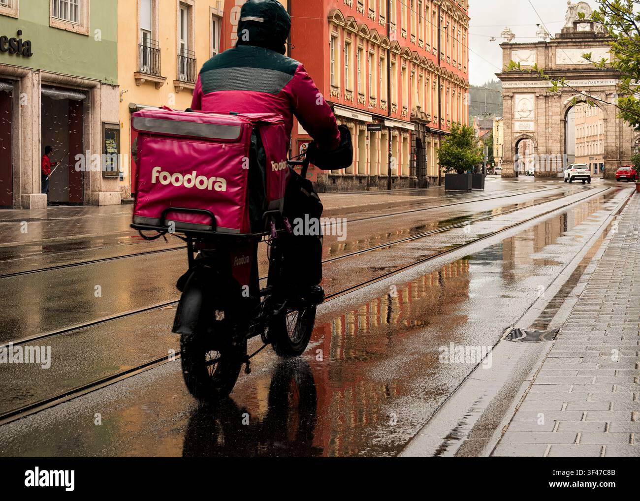 Slancio guidato dalla pioggia mentre un ciclista di consegna spinge attraverso la pioggia urbana riflettente (Inssbruck, Austria: 14-maggio-2026) Foto Stock