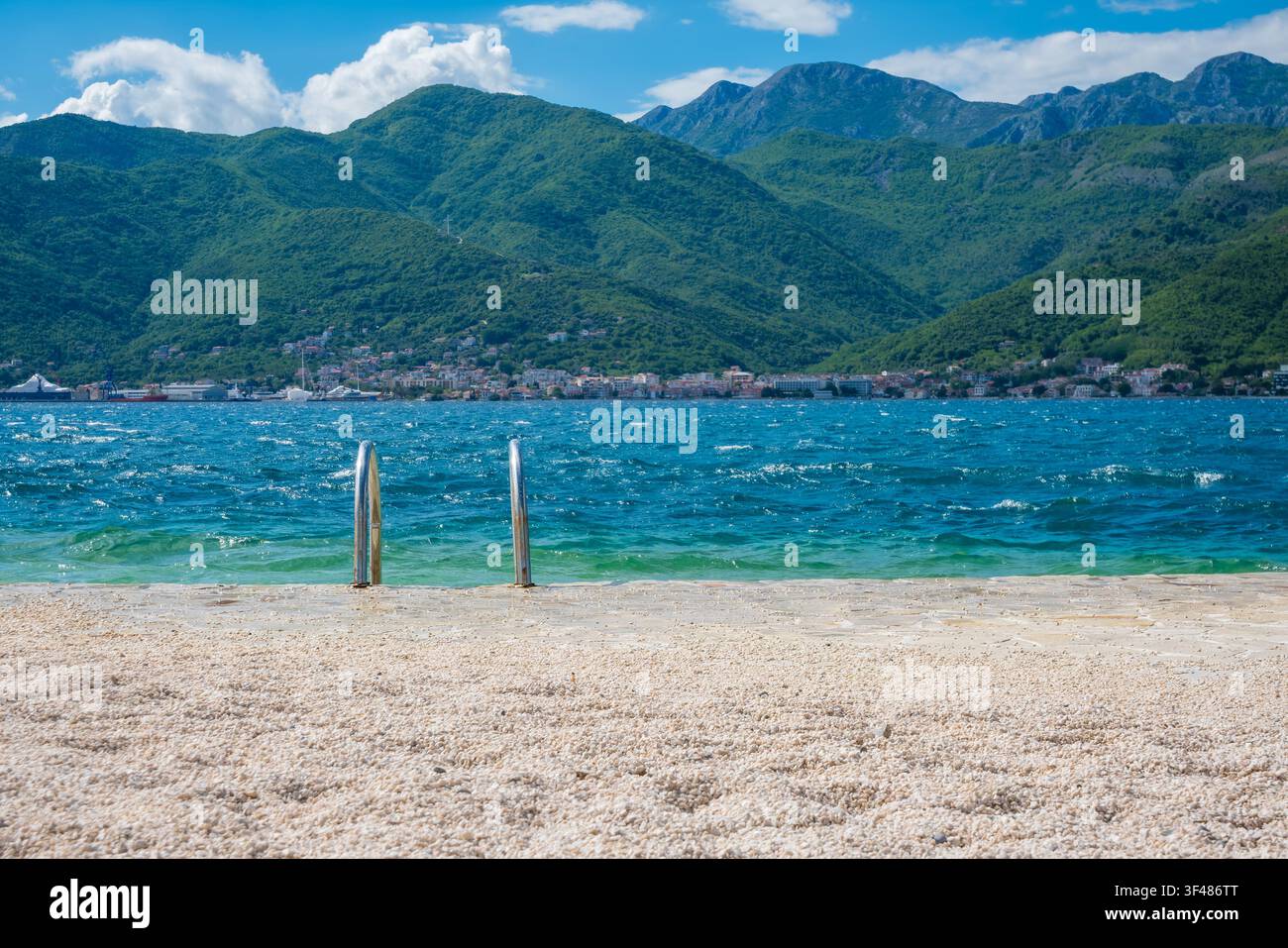 Spiaggia di ciottoli con ringhiere metalliche per l'accesso all'acqua, acqua blu limpida e onde dolci, sfondo di montagne verdi lussureggianti, scena costiera serena. Foto Stock
