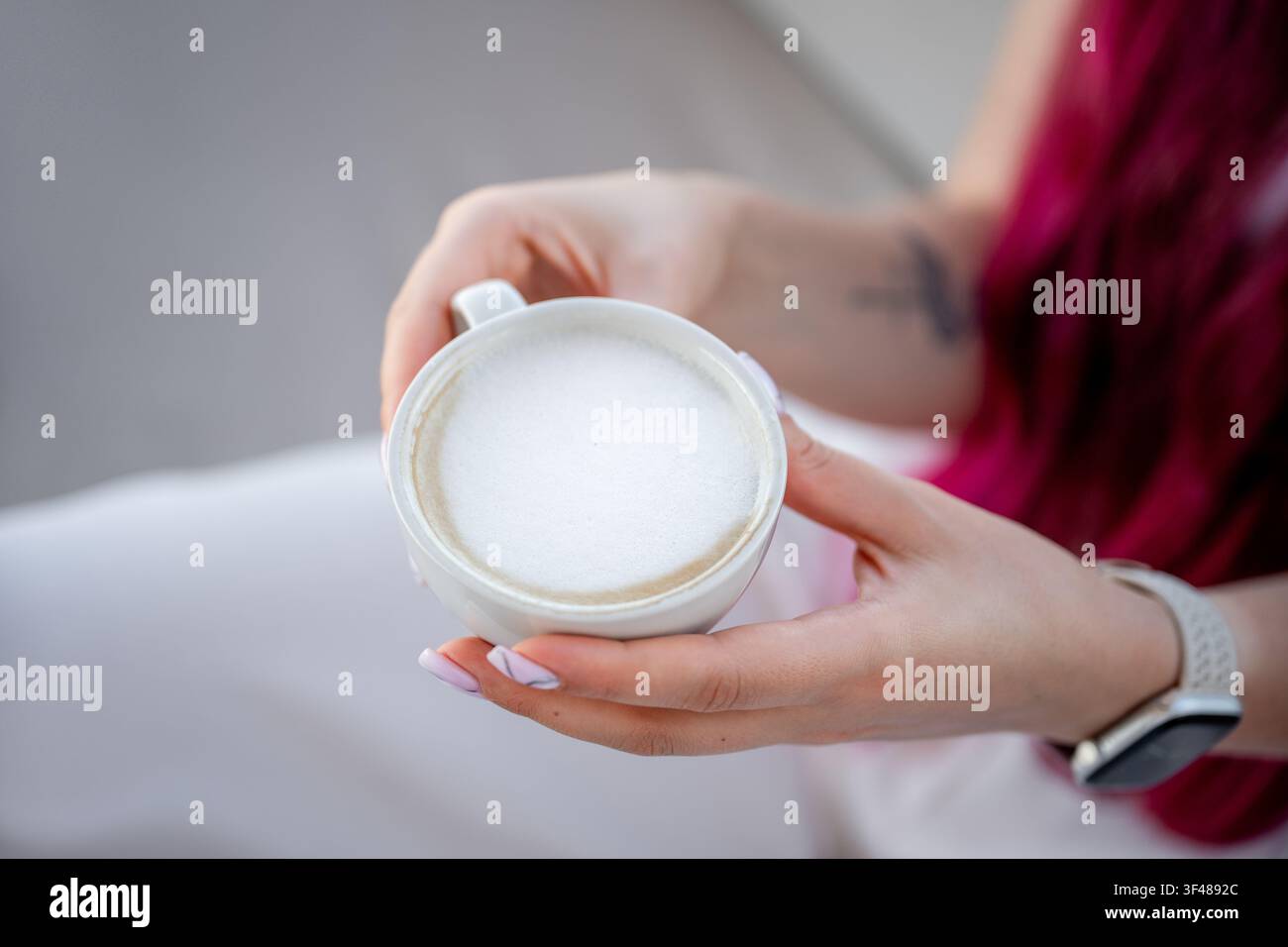 Primo piano delle mani di una donna che tiene una tazza bianca di cappuccino con schiuma di latte cremosa. Momento accogliente in un caffè con luce soffusa, abbigliamento casual e vibrazioni rilassanti Foto Stock