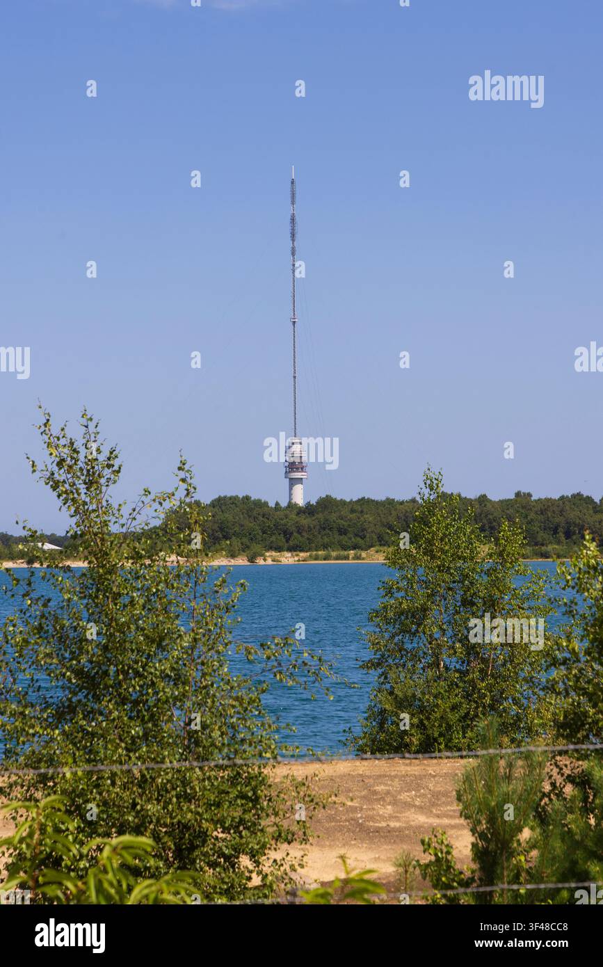 Un'alta torre di trasmissione e un'antenna a Hoogersmilde, Drenthe, Paesi Bassi si ergono sopra una riva boschiva accanto a un lago blu, incorniciati da alberi verdi Foto Stock