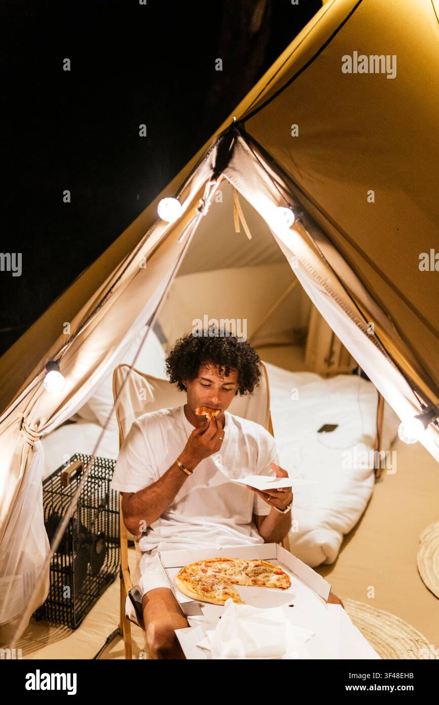Camper maschio solitario siede in una tenda accogliente illuminata da lanterne, mangia pizza e si rilassa durante un tranquillo viaggio in campeggio. Scena di glamping calda in stile interno a nig Foto Stock