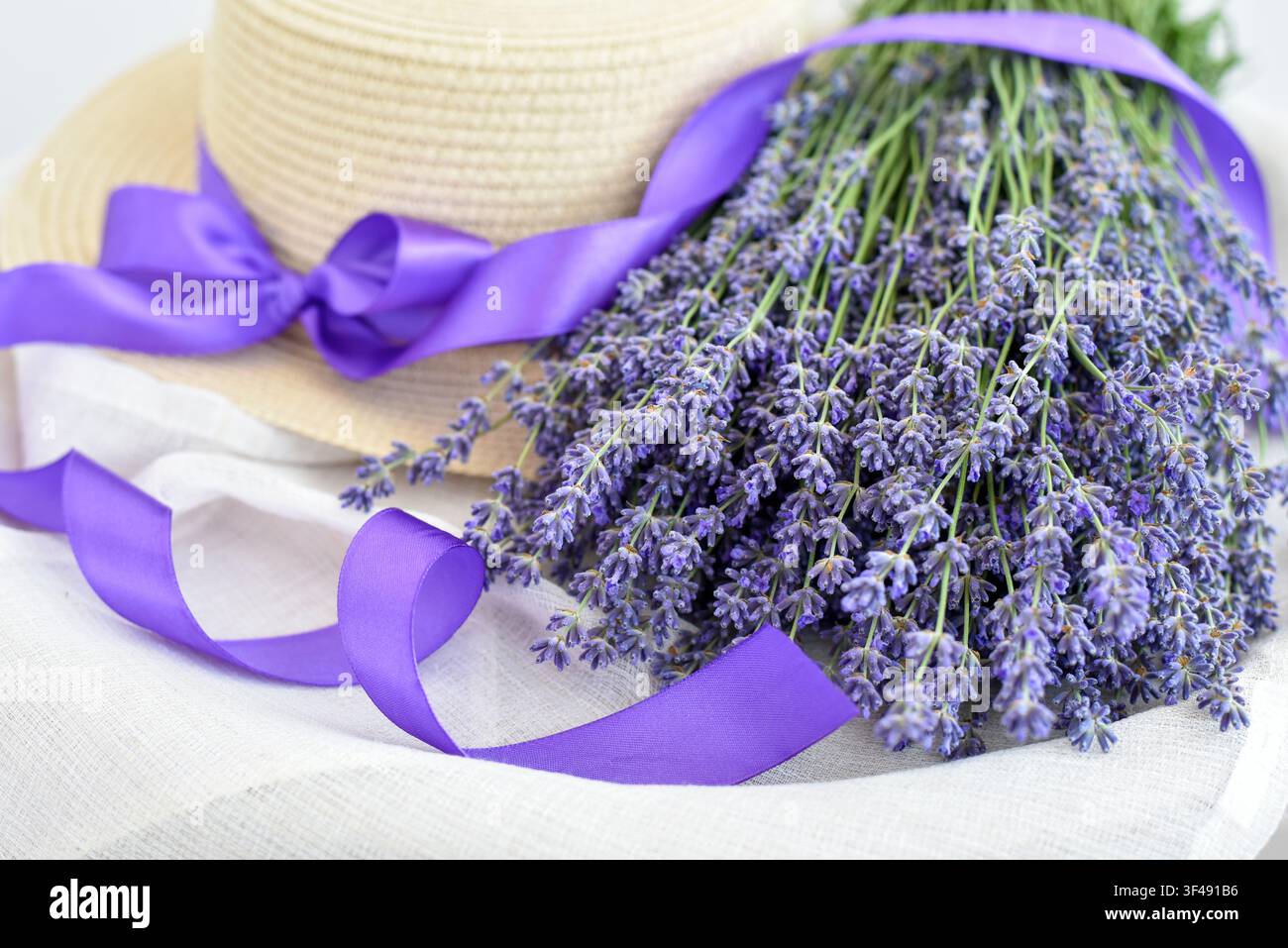Mazzo di lavanda fresca con un cappello di paglia e un nastro viola su tessuto bianco. Sfondo floreale estivo Foto Stock