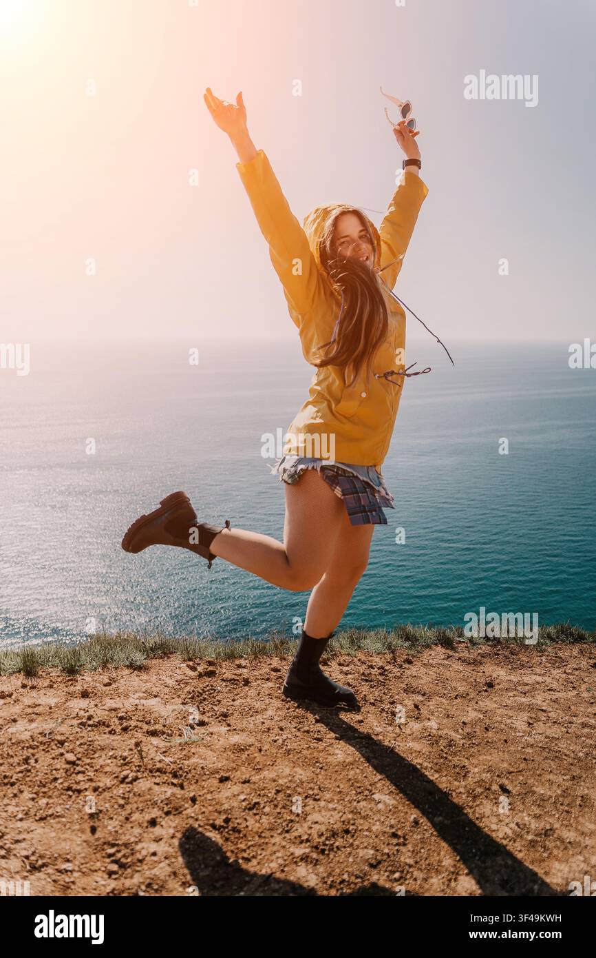 Donna che salta sul bordo della scogliera, donna felice in giacca gialla salta da una scogliera che si affaccia sul mare durante un'escursione soleggiata Foto Stock