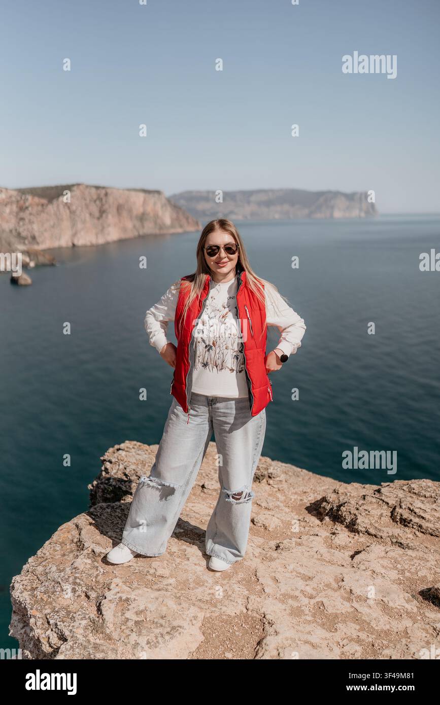 Donna in piedi su una scogliera sull'oceano, su un bordo roccioso panoramico, indossa un giubbotto rosso e si gode la splendida vista sulla costa Foto Stock