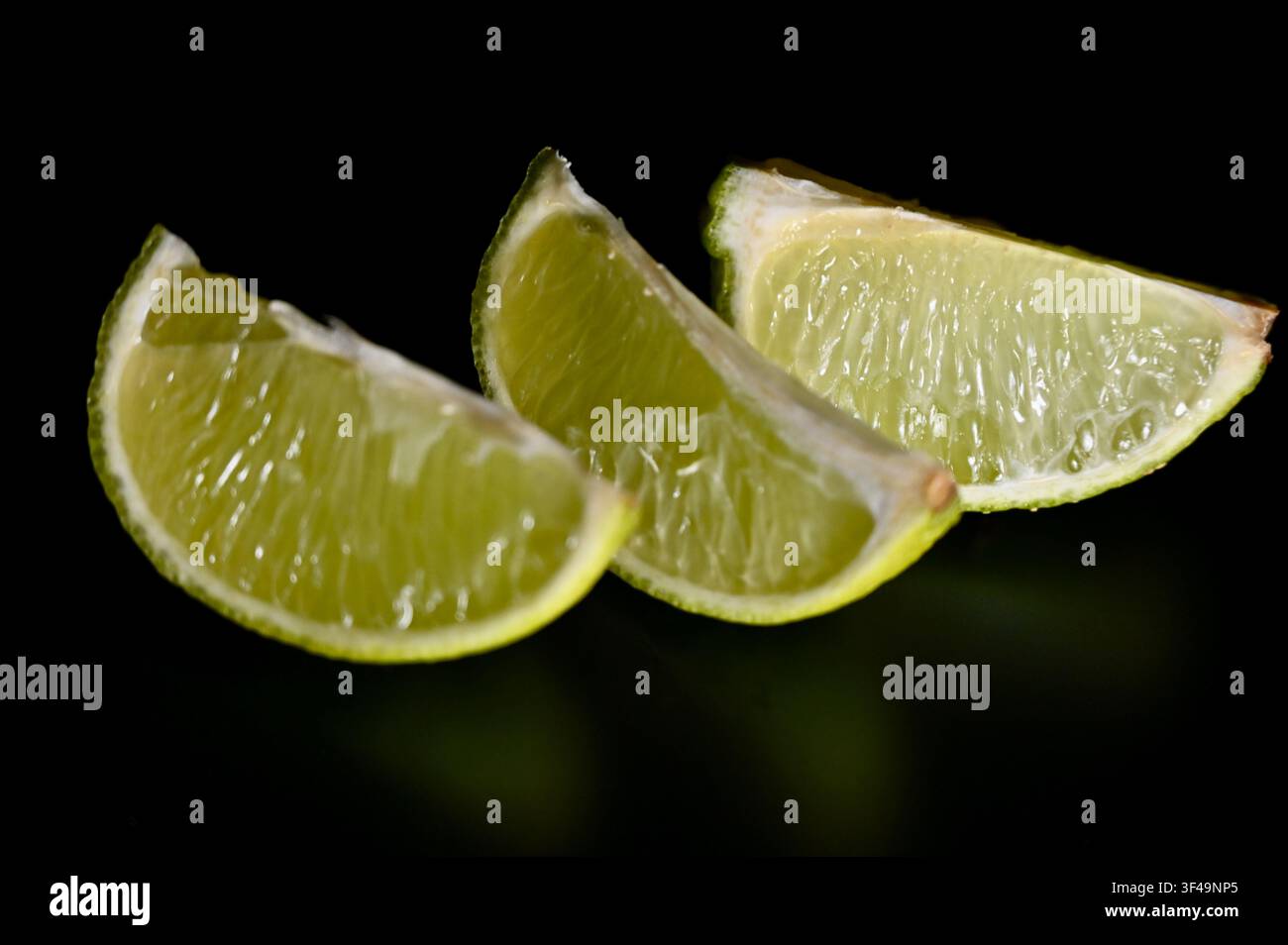 Una foto macro discreta di tre segmenti di limone vivaci allineati in diagonale su una superficie nera come la pece, evidenziando la trama della polpa, la lucentezza naturale e i toni verdi vivaci. Foto Stock