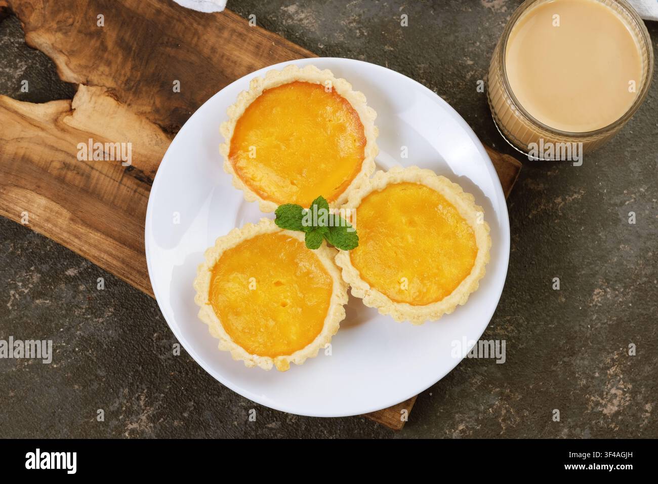 Flat Lay di tre tortine al custard dorate disposte su un piatto bianco con menta fresca e una bevanda cremosa in un contesto rustico da tavolo. Foto Stock