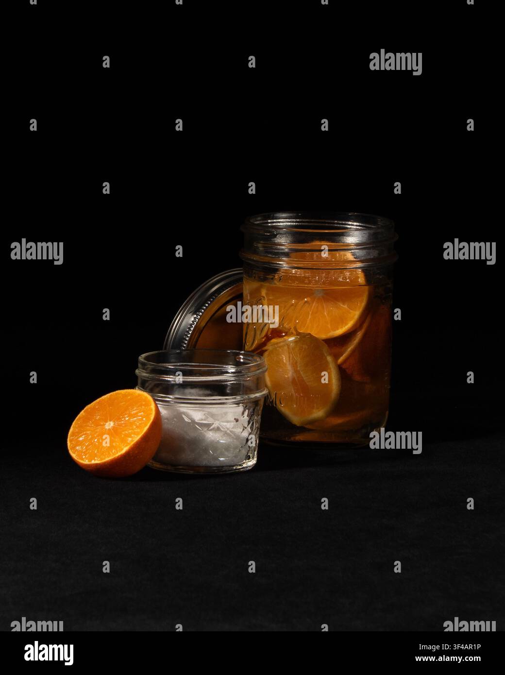 Ingredienti per un miscelatore di cocktail allo sciroppo d'arancia: barattolo Mason pieno di fette di arancia che macerano nell'acqua, piccolo barattolo di zucchero su sfondo di velluto nero. Foto Stock