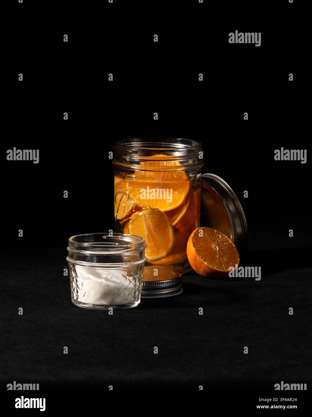 Ingredienti per un miscelatore di cocktail allo sciroppo d'arancia: barattolo Mason pieno di fette di arancia che macerano nell'acqua, piccolo barattolo di zucchero su sfondo di velluto nero. Foto Stock