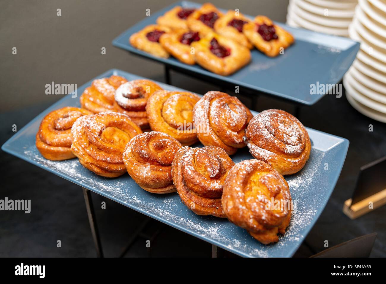 Due vassoi mostrano pasticcini dorati e zuccherati. Il vassoio anteriore contiene panini a spirale con glassa lucida. Il vassoio posteriore mostra croissant ripieni di marmellata. Piatti blu puliti Foto Stock