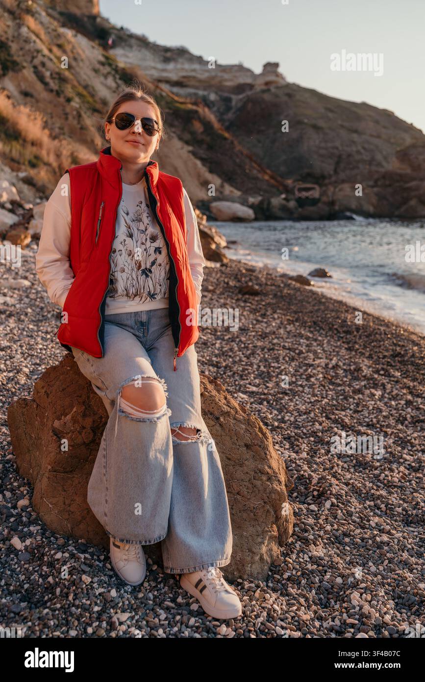 Vestito da spiaggia da donna seduto su una grande roccia lungo la riva di ciottoli durante un tramonto scenografico con montagne Foto Stock