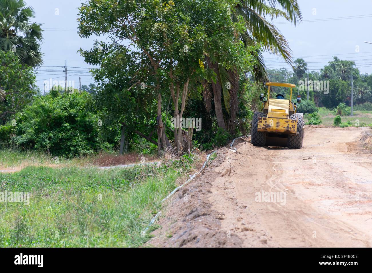 Rullo stradale che compatta una strada sterrata rurale durante la costruzione, circondata da alberi verdi e paesaggio di campagna che evidenzia lo sviluppo delle infrastrutture Foto Stock