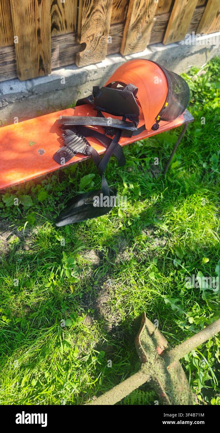Casco di sicurezza con visiera a rete e imbracatura per spalla per tagliasiepi su una panca di legno nel giardino di casa Foto Stock