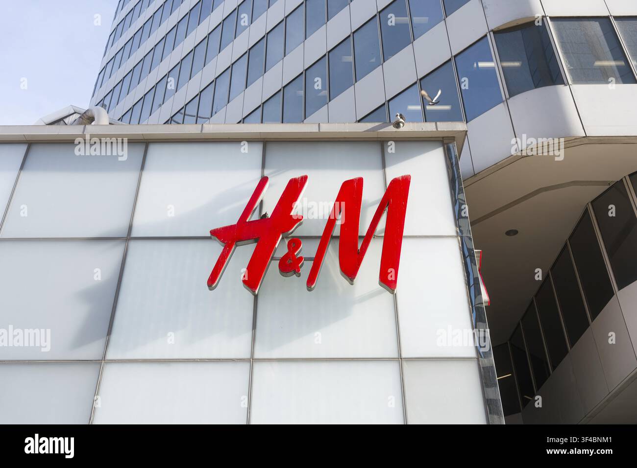 Logo rosso HM su facciata in vetro che trasmette una presenza moderna nel commercio al dettaglio, pannelli riflettenti e griglia bianca, torre di uffici sullo sfondo, centro città soleggiato Foto Stock
