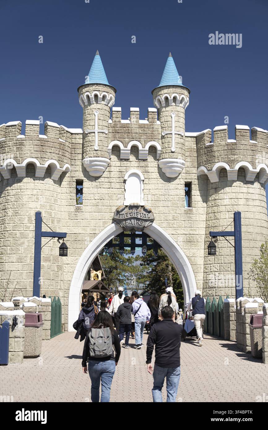 Ingresso del castello nel parco divertimenti con famiglie e visitatori che attraversano un arco di pietra, torri che incorniciano un cielo blu brillante, pavimentazione illuminata dal sole Foto Stock