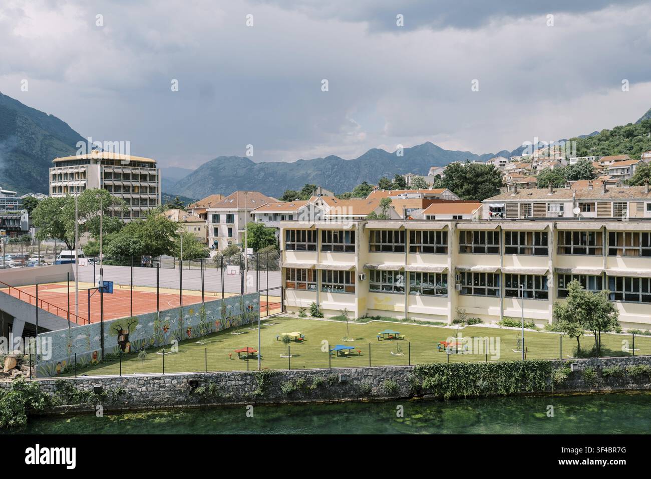 Cortile scolastica di una città di montagna illuminato dal sole, che mostra un campo da tennis, un prato erboso, blocchi di aule a più piani, una recinzione perimetrale, una riva di pietra in primo piano Foto Stock