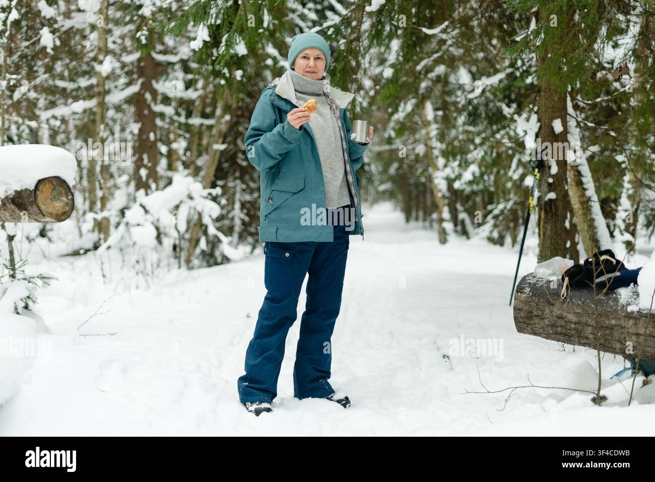 Donna matura che riscalda le mani con una tazza fumante e una thermos in una foresta innevata in inverno. Signora adulta che sorseggia un tè caldo con vapore visibile dalla tazza. Scena accogliente di conforto e calore durante una passeggiata all'aperto nel freddo Foto Stock
