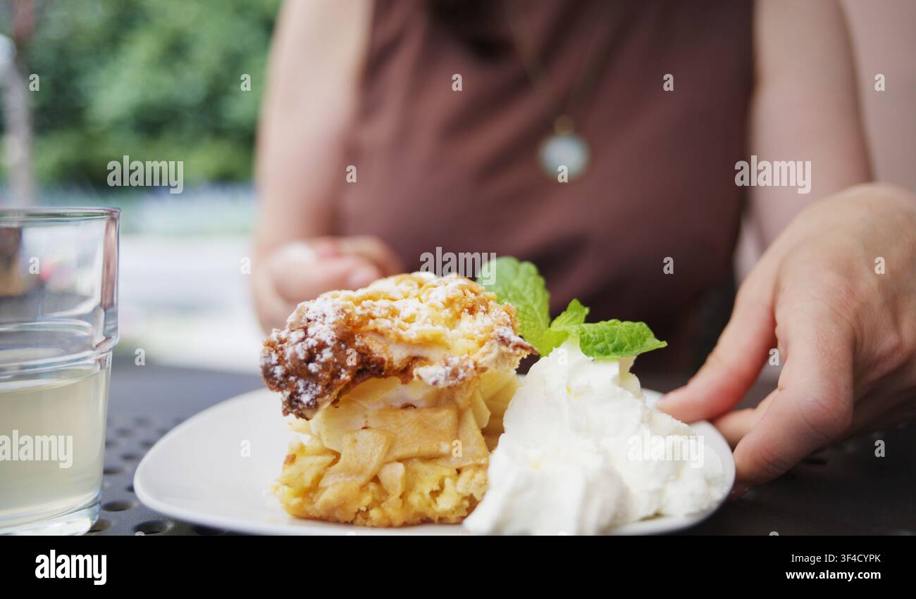 Ragazza si gode una Charlotte di mele con panna e menta, sorseggiando una limonata fredda in un caffè all'aperto ventilato Foto Stock