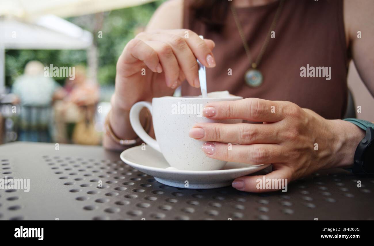 Donna mescola lo zucchero nel caffè a un tavolo di un caffè all'aperto soleggiato, godendosi l'atmosfera calda Foto Stock
