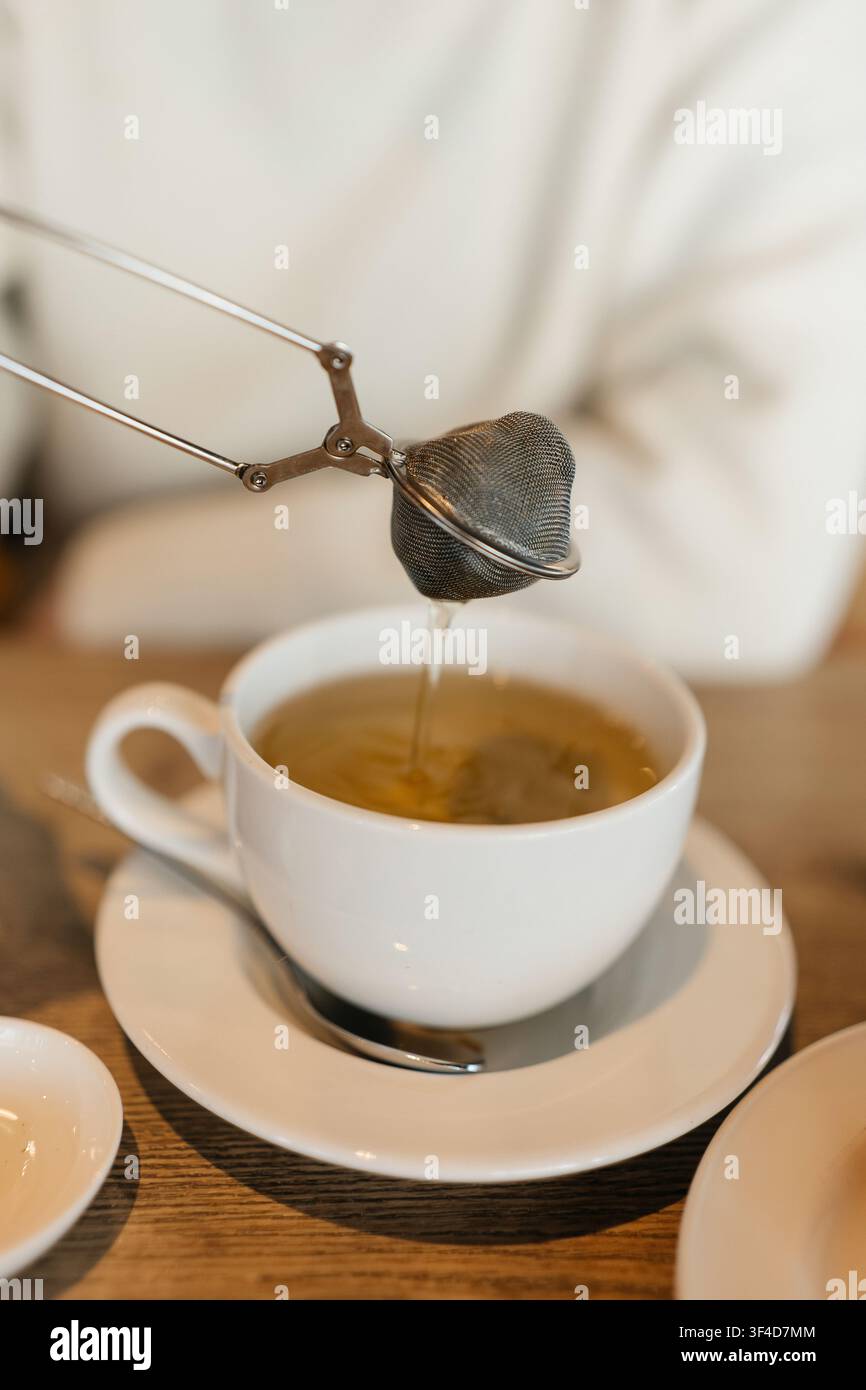 Preparare il tè con un colino di metallo in una tazza bianca su un tavolo di legno Foto Stock
