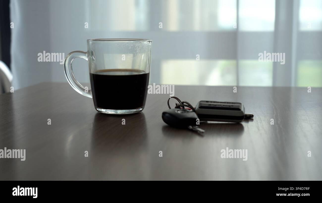 Una vista ravvicinata di un bicchiere di caffè accanto alle chiavi della macchina su un tavolo di legno con una luce naturale soffusa che filtra attraverso tende trasparenti. Foto Stock