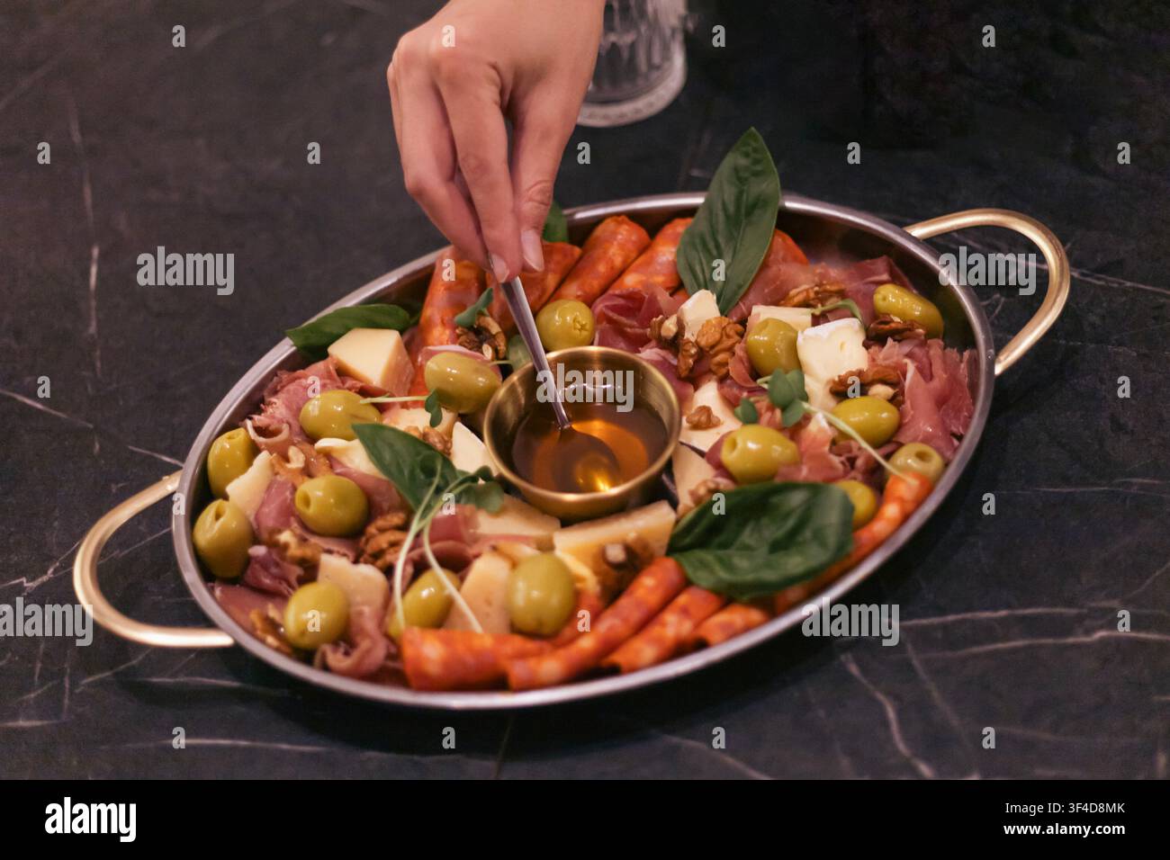 Una persona usa un cucchiaio piccolo per condire con miele un grande vassoio di metallo con carne, formaggi, olive e frutta. Il vassoio è posato su una superficie di marmo scuro Foto Stock