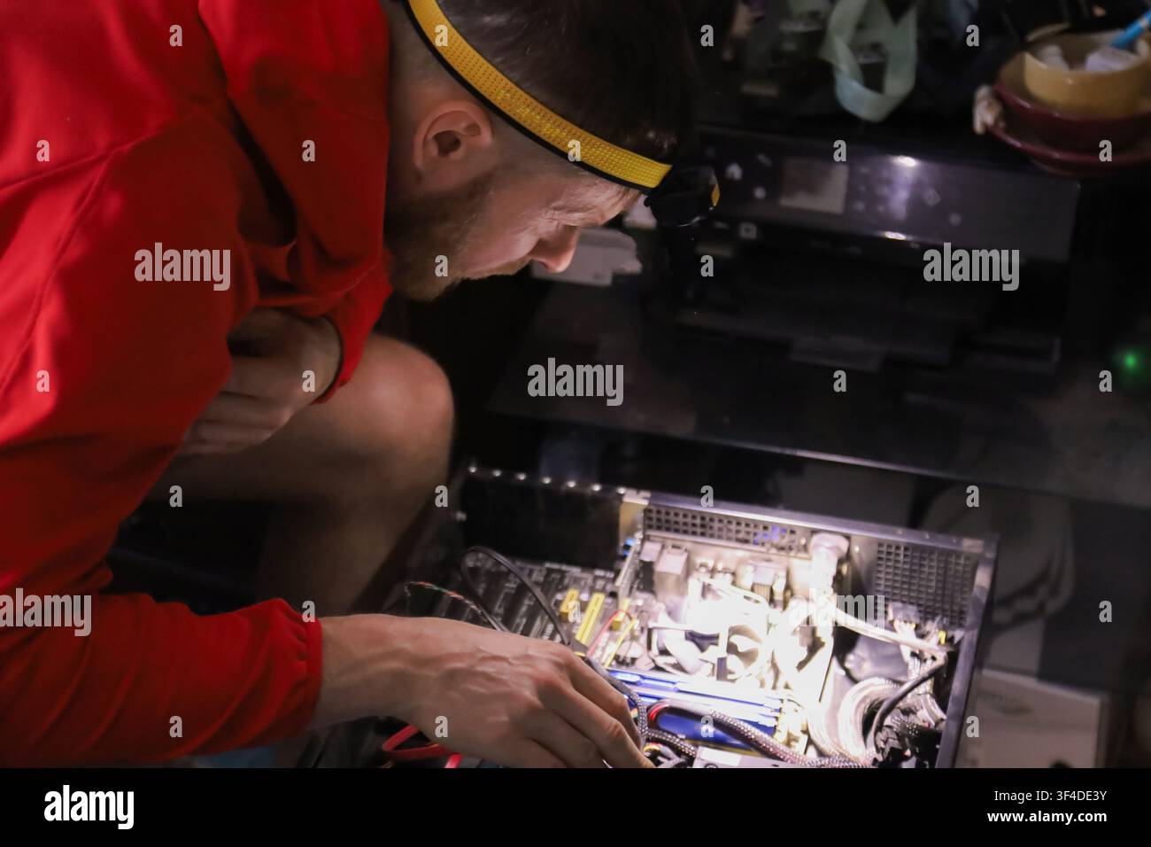 Un uomo con una fascia gialla per capelli e una felpa con cappuccio rossa lavora su una scheda madre del computer. Utilizza strumenti per assemblare componenti elettronici in un ambiente di officina. Foto Stock
