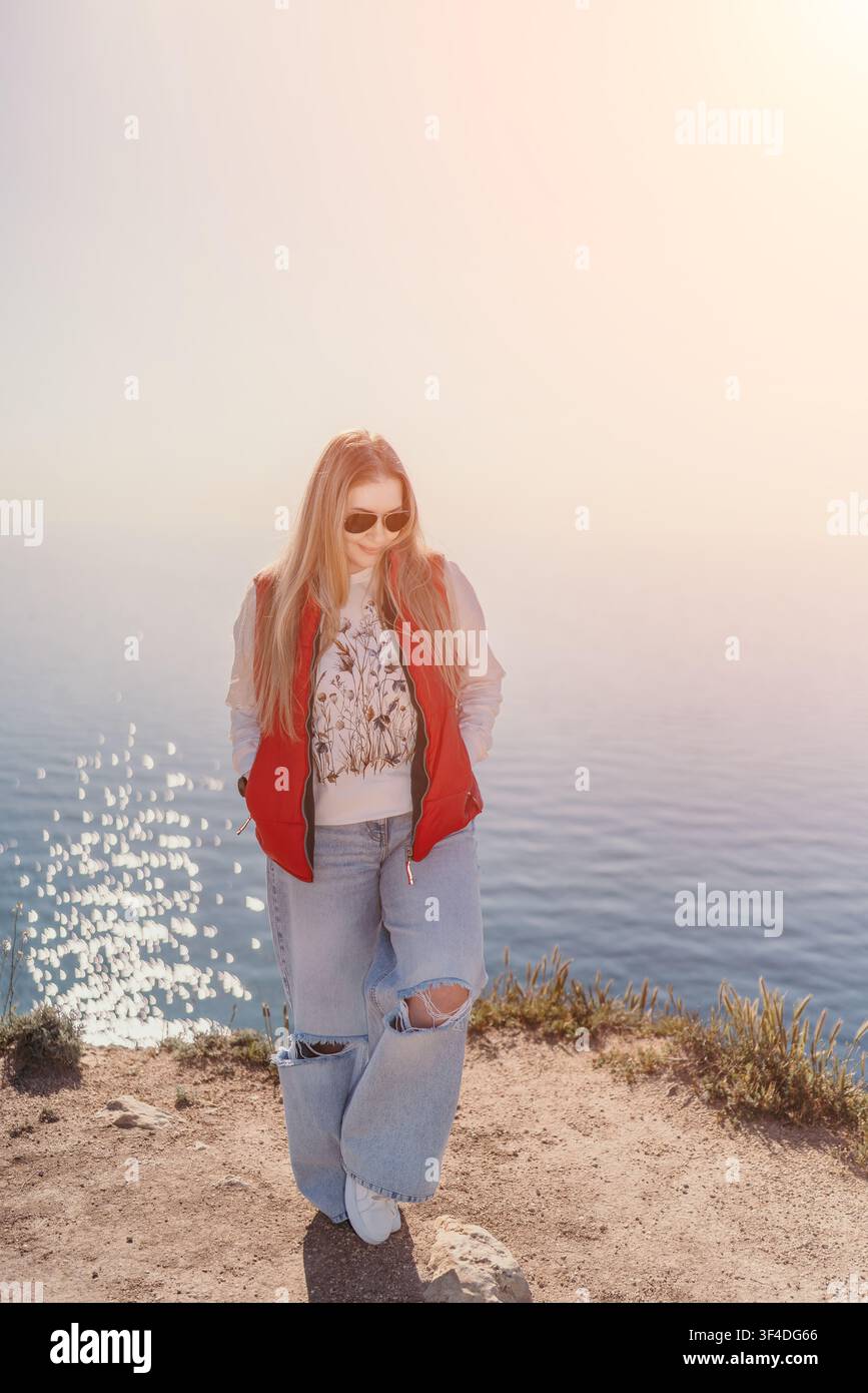 Donna su scogliera sul mare in piedi vicino al bordo dell'oceano che godersi una giornata di sole con giacca rossa e spazio libero Foto Stock