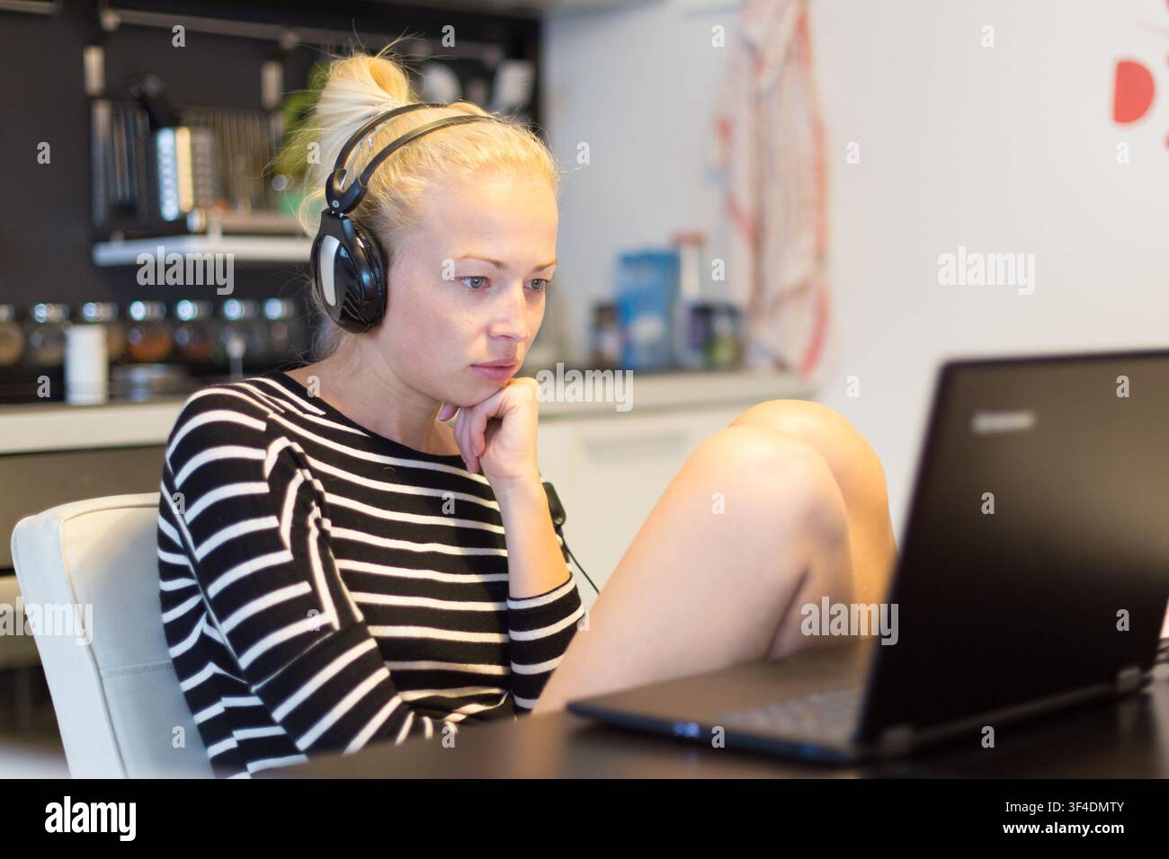Donna informale home abbigliamento lavorano e studiano in remoto dal suo piccolo appartamento a tarda notte. Cucina di casa in background. Grande flessibilità di corsi basati sul web e programmi di studio. Foto Stock