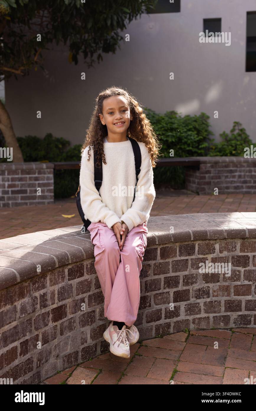 Bambina seduta su panca curva in mattoni nel cortile, indossa un maglione in pile color crema e uno zaino Foto Stock