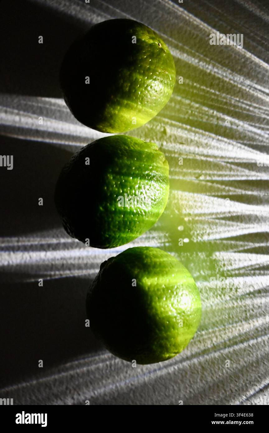 Un primo piano dall'alto di tre lime verdi freschi interi allineati su un vassoio di vetro a fantasia, evidenziati da una forte luce direzionale che crea raggi di luce drammatici e motivi ombra. Foto Stock
