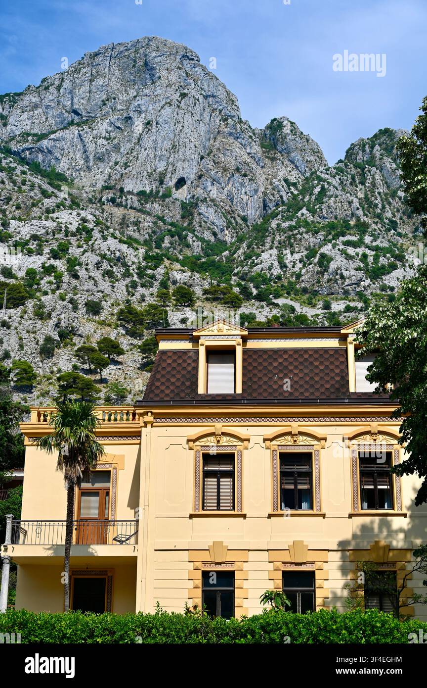 Un colpo all'aperto che mostra una classica villa europea gialla con un tetto a mansarda marrone e un balcone, situata di fronte a imponenti scogliere rocciose grigie sotto un cielo blu terso, circondata da palme e fogliame verde. Foto Stock