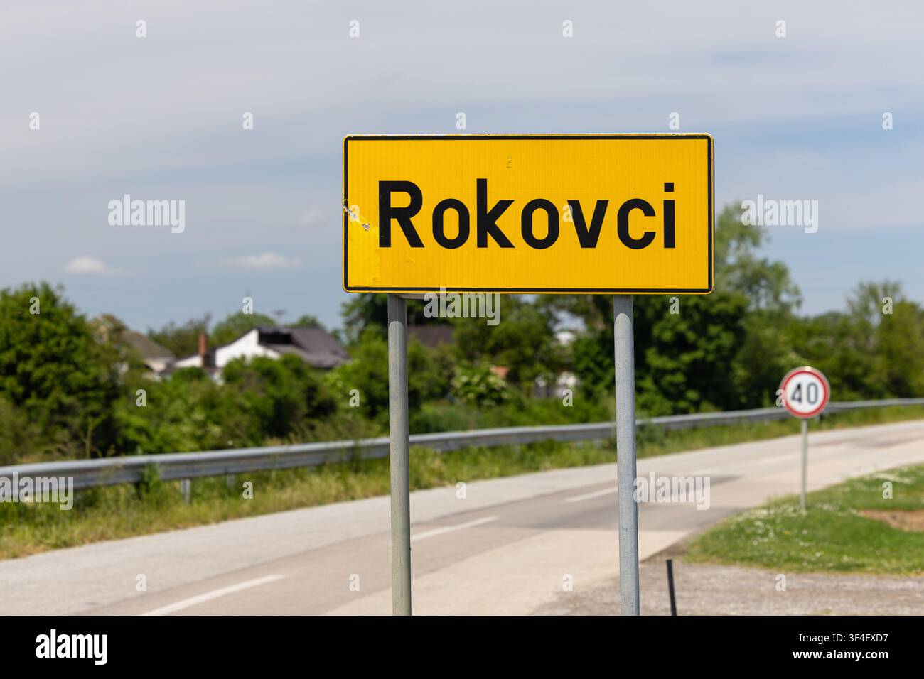 Segnale stradale del villaggio di Rokovci montato su pali metallici accanto a una strada asfaltata rurale con un cartello di limite di velocità a 40 km/h, guardrail e campagna verde sullo sfondo Foto Stock