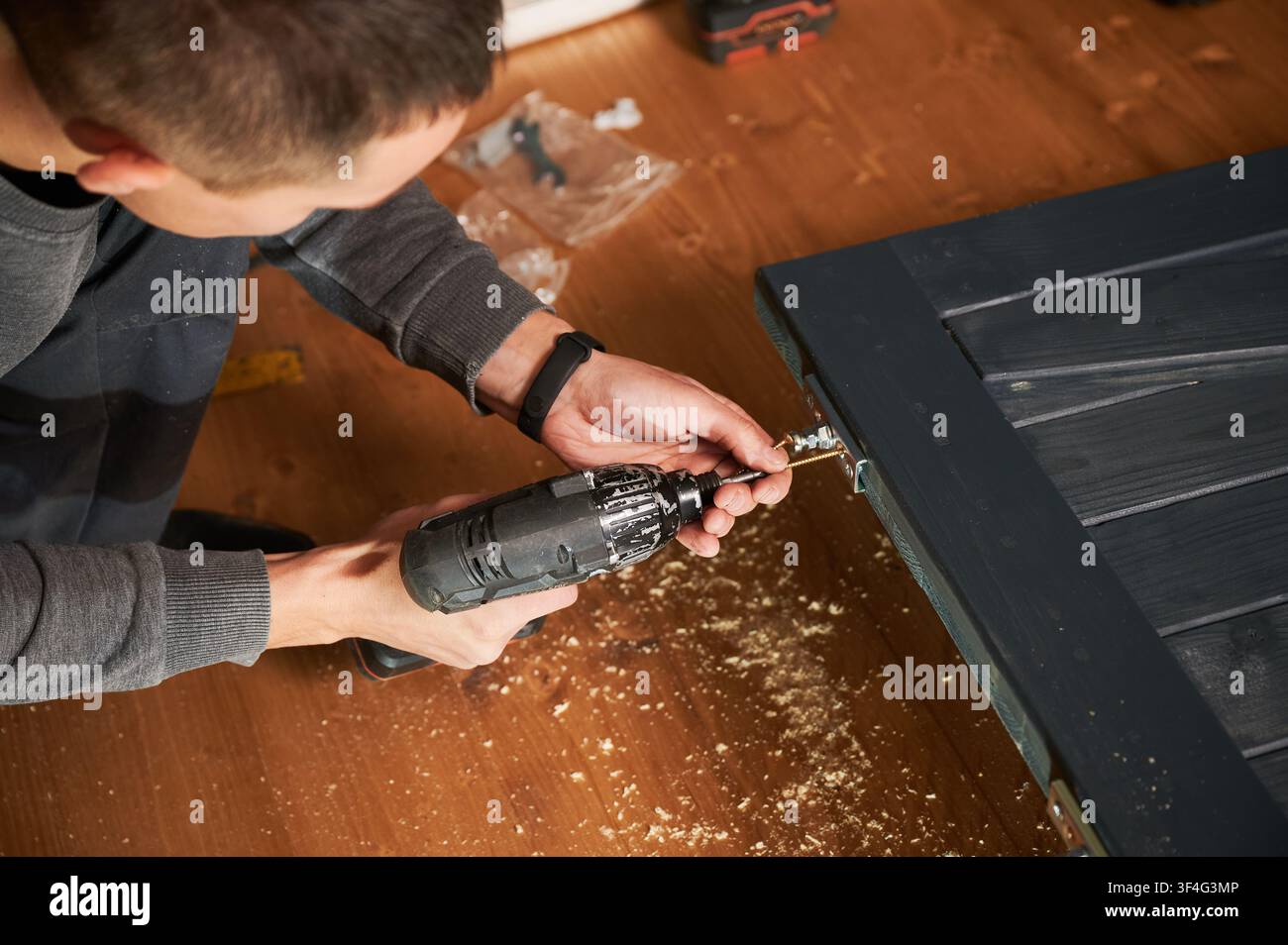 Falegname che avvitata una vite in una piastra metallica su una superficie di legno con un trapano, primo piano del lavoro dettagliato. Officina interna con trucioli e attrezzature, concetto di assemblaggio e fissaggio di mobili Foto Stock