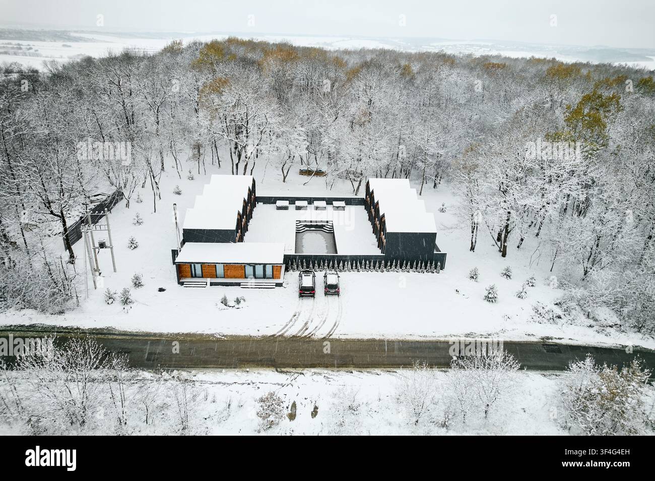 Case moderne di lusso situate nella campagna invernale. Prospettiva a dronata elevata di edifici geometrici con cortile centrale e piscina, auto parcheggiate lungo la strada, sfondo boscoso, concetto di vita esclusivo Foto Stock